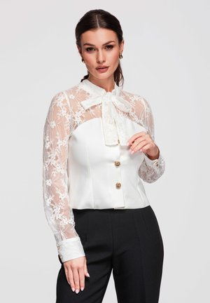 Femme aux cheveux foncés dans un chemisier en dentelle blanche avec un nœud et des boutons dorés, associée à un pantalon noir taille haute, posant sur fond blanc.