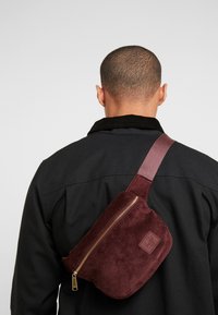 Mężczyzna widziany z tyłu, ubrany w czarną kurtkę i bordową torbę na ramię z sztruksu, z złotym zamkiem z tyłu.