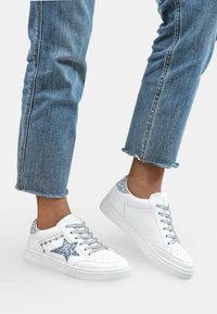 Baskets blanches avec des accents en paillettes argentées, motif d'étoile, clous décoratifs, semelle texturée et lacets gris, associées à un jean bleu à bord effiloché.