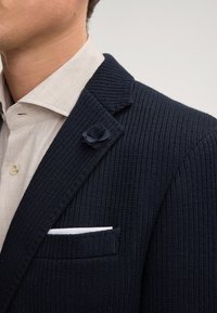 Blazer a righe blu navy in tessuto testurizzato, con un accento floreale sul bavero e un fazzoletto bianco nella tasca. Camicia color chiaro con colletto sotto.