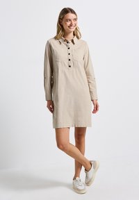 Robe chemise beige avec des manches longues, patte de boutonnage à l'avant, poche poitrine et coupe décontractée. Portée avec des baskets blanches et beiges. Texture de tissu lisse.