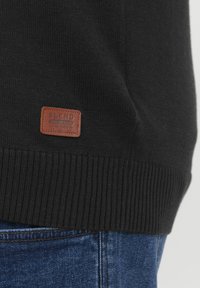 Pull en tricot noir avec bord-côte. Présente une étiquette en cuir marron rectangulaire embossée avec "BLEND" et les détails de l'établissement.