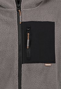 Grijze fleece jack met een zwart ritsvak, met een gestructureerde oppervlak en een bruin logo label. Rits met een tan kleurige koord en slider detail.