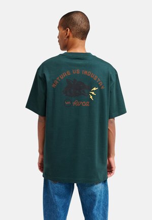 Uomo che indossa una t-shirt verde scuro con grafica di un lupo e testo "Nature US Industry" e jeans blu, in piedi di spalle su sfondo bianco.
