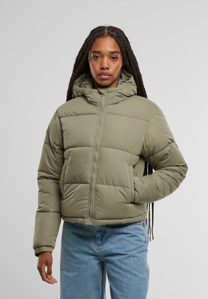 Doudoune vert olive avec un col montant, design matelassé, fermeture éclair à l'avant et poignets élastiques. Porte sur un jean bleu clair.