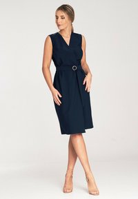 Granatowa sukienka bez rękawów z dekoltem w serek, przewiązana w talii złotym pierścionkiem, do kolan, o gładkiej fakturze materiału.