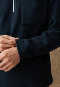 Námornícky fleecový top s golierom na zips do štvrťky, s vreckom na prsiach a manžetami na dlhých rukávoch. Materiál vyzerá mäkký a textúrovaný.