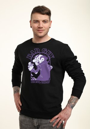 Mann mit kurzen Haaren, Ohrpiercings und Tätowierungen, der einen schwarzen Pullover mit lila Cartoon und dem Text „Bad Guy Be Evil Today“ trägt.