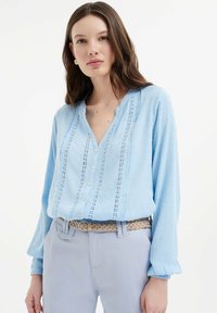 Blusa azzurro chiaro con maniche lunghe, chiusura con bottoni, dettagli in pizzo intricato e pois testurizzati, abbinata a pantaloni chiari e una cintura intrecciata.