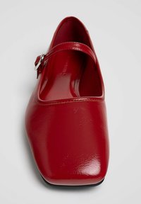 Chaussure Mary Jane rouge brillante à bout carré avec bride à boucle argentée sur fond gris clair.