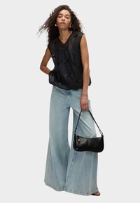 Top senza maniche nero con fiocchi, in materiale leggero e trasparente, abbinato a jeans blu chiaro a gamba larga, mentre tiene una piccola borsa in pelle nera.