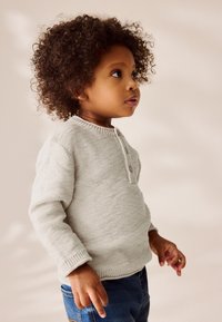 Maglione grigio chiaro lavorato a maglia con scollo rotondo, dettagli con bottoni e polsini a coste, abbinato a jeans di denim blu. Bambino in piedi di profilo.