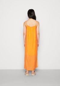 Robe maxi orange avec des fines bretelles et un col volanté. Tissu léger avec une silhouette fluide, fentes sur les côtés, portée sur des chaussures beiges.