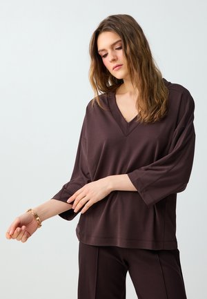 Junge Frau richtet den Ärmel eines locker sitzenden dunkelbraunen V-Ausschnitt-Tops, trägt passende Hosen und ein goldenes Armband, vor hellem, einfachem Hintergrund.