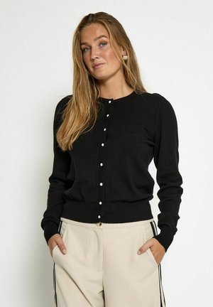 Black knitted cardigan with long sleeves, round neck, and white button details; paired with beige pants featuring side stripes.