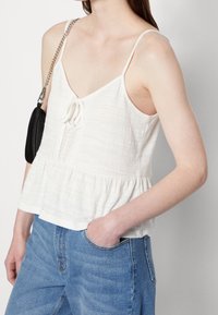 Débardeur blanc texturé avec un ourlet peplum, bord en dentelle et un lien avant. Associé à un jean bleu taille haute. Sac noir à chaîne.