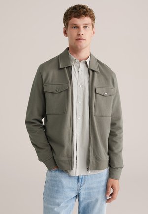 Olive green, zip-up jacket with two chest pockets, ribbed cuffs, and collar. Worn over a striped, collared shirt and light blue jeans.