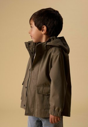 Ragazzo giovane di profilo che indossa una giacca con cappuccio verde e jeans azzurri chiari su sfondo beige.