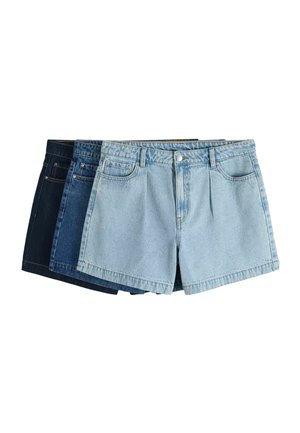 Frau mit kurzen Haaren, die einen schwarzen schulterfreien Pullover und hellblaue hochgeschnittene Jeansshorts vor einem einfarbigen Hintergrund trägt.