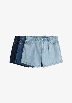 Femeie cu păr scurt care poartă un pulover negru cu umerii goi și pantaloni scurți din denim bleu deschis cu talie înaltă pe un fundal simplu.