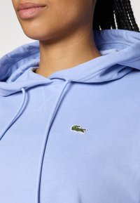 Hanorac bleu deschis, realizat dintr-un material moale, cu o buzunar tip cangaroo și un mic logo verde în formă de crocodil pe partea stângă a pieptului.