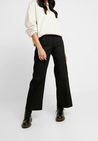 Czarne spodnie o szerokich nogawkach z wysokim stanem, wykonane z teksturowanej tkaniny, w połączeniu z kremową, oversize'ową bluzą i czarnymi, sznurowanymi botkami.