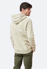 Sudadera beige con capucha ajustable, textura suave y un corte relajado, acompañada de pantalones verde oliva. Costuras visibles y puños acanalados.