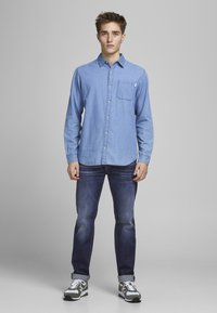 Cămașă de chambray albastru deschis cu un buzunar pe piept, cu nasturi pe față, mâneci lungi, asortată cu blugi albastru închis și adidași verzi cu alb.