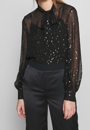 Frau trägt eine transparente schwarze Bluse mit goldenen Polka Dots, eine Schleife als Krawatte und schwarze Satin-Hochbundhose.