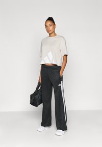 Znižana bež majica z belo grafiko, kombinirana s črnimi trenirkami z belimi stranskimi črtami in logotipom Adidas, držanje črne torbe.