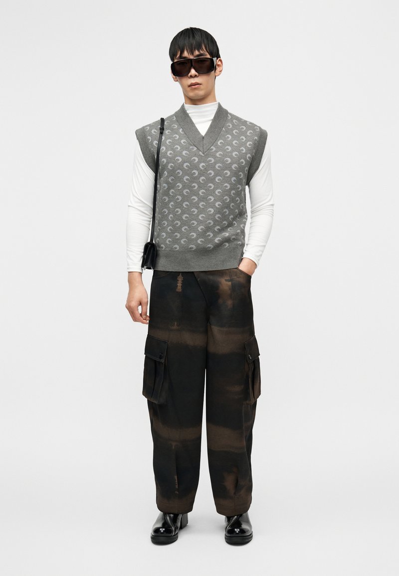 Γκρι πλεκτό γιλέκο με διακοσμητικά σχέδια πάνω α πό λευκό πουκάμισο μακρυμάνικο, συνδυασμένο με σκούρο παντελόνι cargo με τσέπες, ολοκληρωμένο με μαύρα παπούτσια.