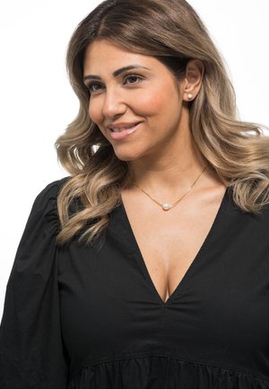 Black v-neck dress with puff sleeves, featuring a gathered waist. Gold necklace with a single white pearl accent. Wavy, shoulder-length hair.