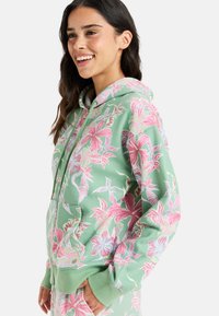 Felpa verde con zip e stampa floreale nei toni del rosa e dell'azzurro. Presenta un cappuccio foderato, tasche frontali e polsini a coste. Realizzata in tessuto morbido.