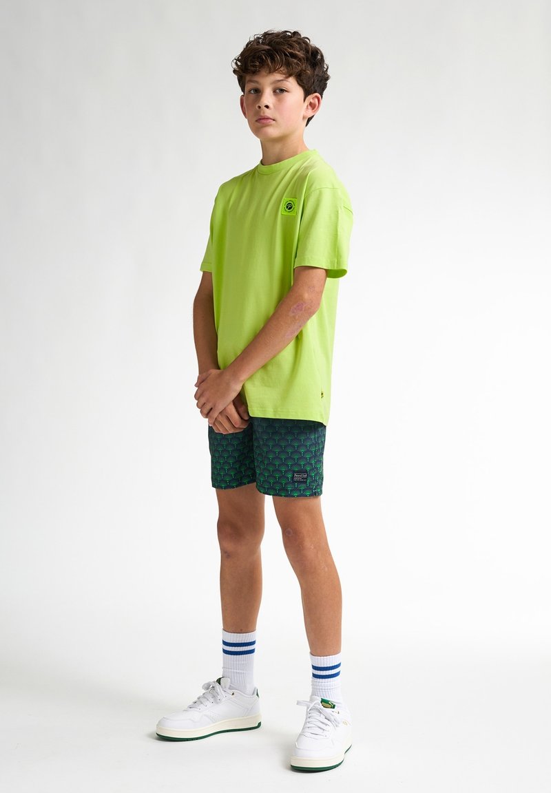 Neon groene katoenen t-shirt met korte mouwen en een logo op de borst, gecombineerd met gestreepte marineblauwe shorts met palmbomenmotieven. Witte sneakers en gestreepte sokken.