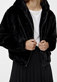 Black faux fur bomber jacket with a hood, zippered front, and elasticized cuffs. Worn over a white t-shirt and a pleated black skirt.