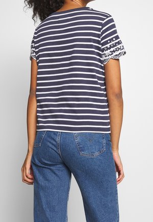 Femme portant un t-shirt à manches courtes rayé bleu marine et blanc avec un bord en dentelle florale sur les manches et un jean bleu, vue de dos.