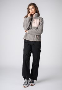 Grau Fleece-Jacke mit pinkem Einsatz, frontzem und hohem Kragen. Kombiniert mit schwarzen Cargo-Hosen und schwarz-weißen Sneakers.