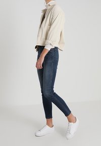Beige corduroy cropped jacket with a pocket, paired with dark blue slim-fit jeans and white sneakers. Clean lines and simple design.