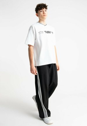 Młoda osoba stojąca, ubrana w białą, dużą koszulkę z czarnym napisem oraz czarne spodnie dresowe z białymi paskami, białe trampki.