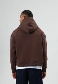Bruine pullover hoodie met een grote capuchon, een relaxte pasvorm en geribbelde manchetknopen; gedragen over een wit shirt, gecombineerd met donkerblauwe denim jeans.