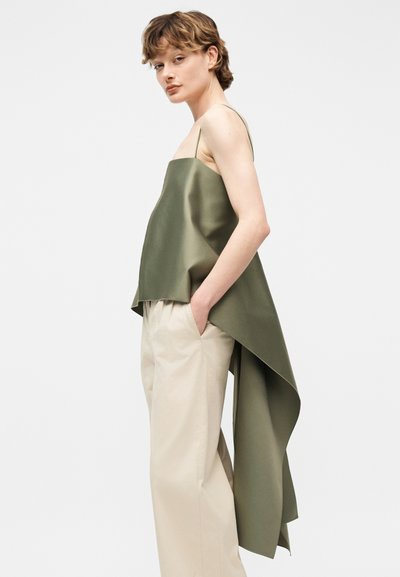 Γυναίκα που φοράει πράσινο ελιάς ασύμμετρο αμάνικο τοπ με μακρύ πέπλο στο πίσω μέρος και μπεζ παντελόνι φαρδιάς γραμμής, στέκεται με τα χέρια στις τσέπες.