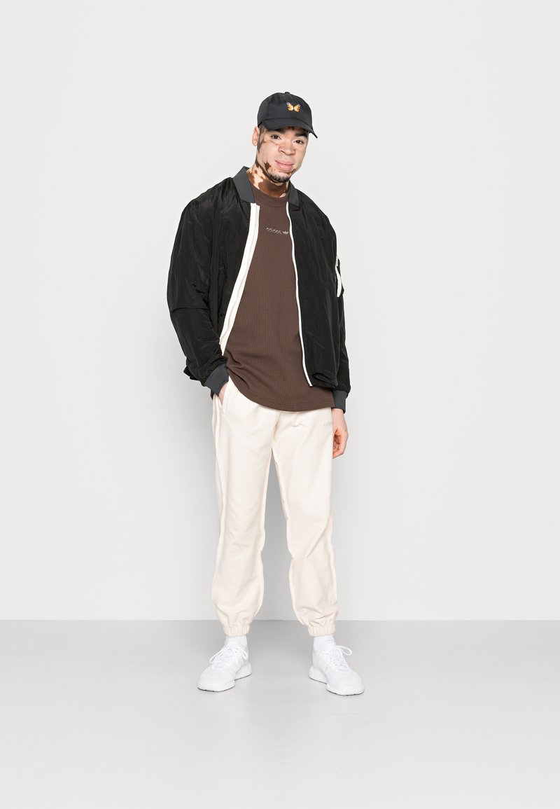 Chaqueta bomber negra con cremallera blanca, camiseta de canalé marrón, pantalones jogger crema y zapatillas blancas; gorra negra con un logo de mariposa.