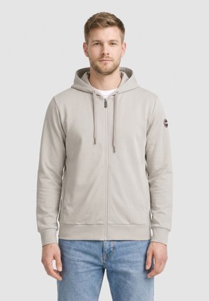 Man draagt een lichtgrijze zip-up hoodie met trekkoorden en blauwe spijkerbroek, staand tegen een neutrale achtergrond.