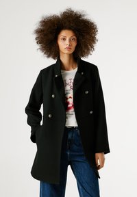 Melna divrindu jaka ar sudraba pogām, ar augstu apkakli un gludu tekstūru, stilizēta virs baltas grafiskas T-krekla un zilajiem džinsiem.