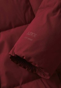 Vínová bunda s prošívanou texturou, vybavená elastickými manžetami a potiskem značky "JUXX EST. 2020" na rukávu.