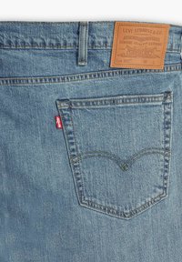 Valaistusyksinkertainen sininen farkut, joissa on vaaleanruskea nahkapatchi vyötäröllä, takatasku punaisella Levi's-tunnuksella sekä erottuvat kaarevat ompelutiedot.