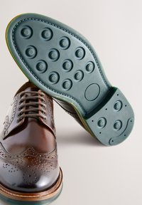 Brun lædersko med brogue-detaljer, der har en rundet tå og en grøn, gummisål med cirkulære grebsmønstre.