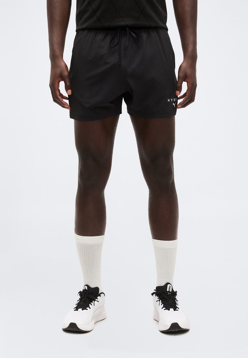 Uomo che indossa pantaloncini atletici neri, calzini bianchi a metà polpaccio e scarpe da ginnastica bianche con lacci neri, in piedi contro uno sfondo chiaro uniforme.