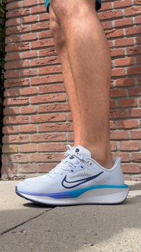 Nike löparsko i vitt med blå accenter, texturerad mesh-överkonstruktion, vadderad häl och mönstrad sula, placerad mot en tegelvägg.