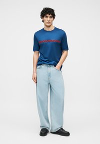 Νέος άνδρας που φοράει μπλε μπλουζάκι Emporio Armani, ανοιχτό μπλε τζιν με φαρδιά μπατζάκια και μαύρα παπούτσια, στέκεται μπροστά σε μονόχρωμο φόντο.